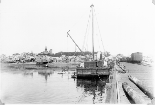 Railroad wharf with view of High Pole Hill in the background. Pictured is a barge with a crane that lifted the granite blocks off of ships from Stonington, Maine. The blocks were loaded onto carts and transported to the base of High Pole Hill. There the granite was loaded onto motorized rail carts, which carried the blocks up the hill to the construction site. Circa Spring 1907