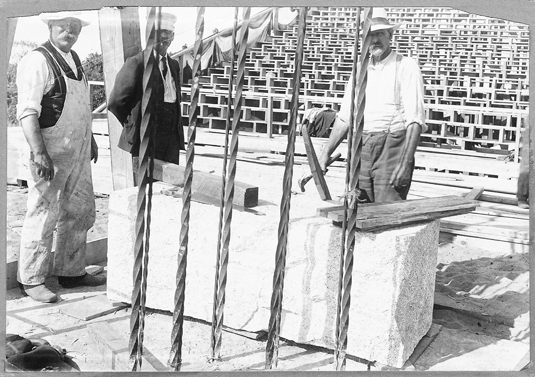 Three work men at foundation of the Pilgrim Monument, preparing the granite cornerstone to be laid by President Roosevelt, with newly constructed grandstand in the background. Circa August 1907.